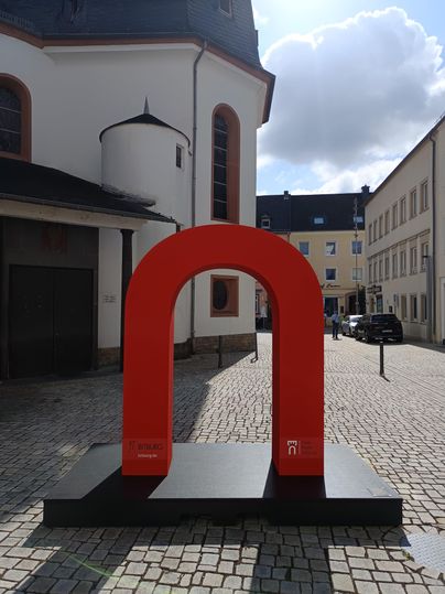 Een grote, rode sculptuur in de vorm van de letter "n" staat op een plein. Op de achtergrond zijn historische gebouwen en een heldere lucht te zien.