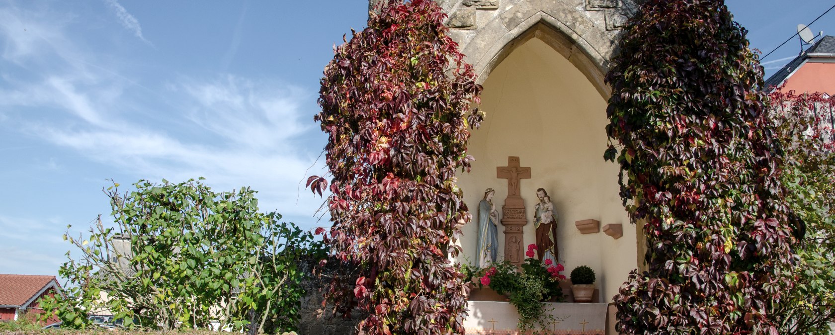 Eine kleine Kapelle in Wolsfeld, umgeben von roten Kletterpflanzen. Im Inneren sind religiöse Statuen zu sehen. Der Himmel ist klar und blau., © Tourist-Information Bitburger Land