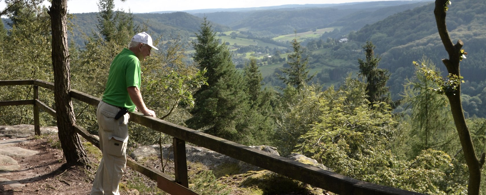 A man in green clothing stands at a railing and looks out over a wooded landscape with hills and valleys., © Lauschtour.de