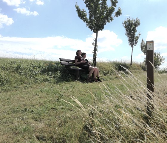 Zwei Personen sitzen auf einer Bank in einer grünen Landschaft. Der Himmel ist blau mit einigen Wolken. Ein Wegweiser steht rechts im Bild., © Tourist-Information Bitburger Land