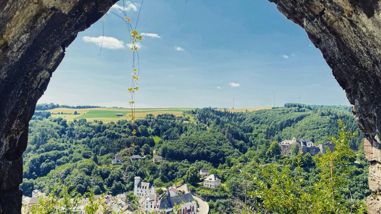 Ein Blick durch einen alten Bogen auf eine grüne Landschaft. Im Hintergrund sind Hügel und kleine Gebäude sichtbar, unter einem klaren blauen Himmel.