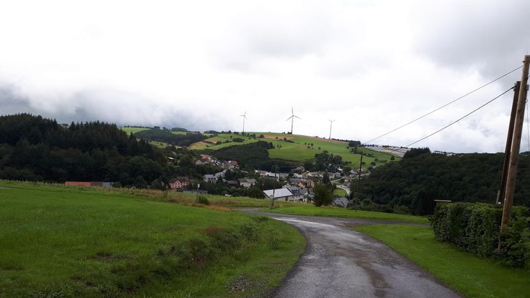 Un paysage tranquille avec des collines douces et un chemin étroit. En arrière-plan, on voit des éoliennes et un petit hameau.