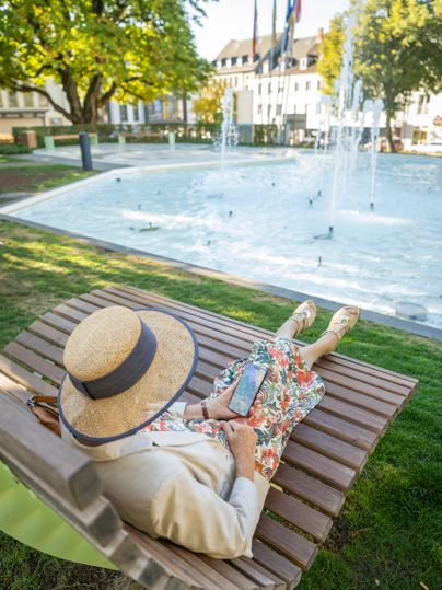 Eine Frau sitzt entspannt auf einer Holzliege und schaut auf ihr Smartphone. Im Hintergrund ist ein Springbrunnen und grüne Bäume zu sehen.