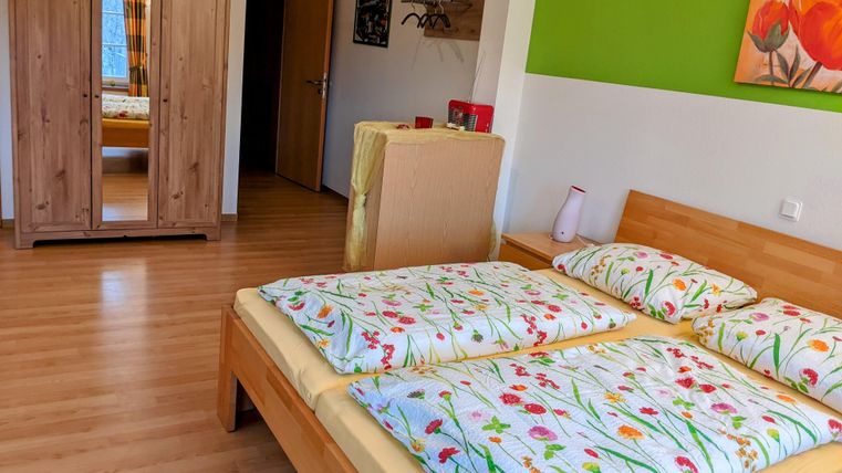 Ein gemütliches Schlafzimmer mit zwei Betten und bunten Bettbezügen. Im Hintergrund steht ein Kleiderschrank und eine Wand mit einem grünen Akzent.