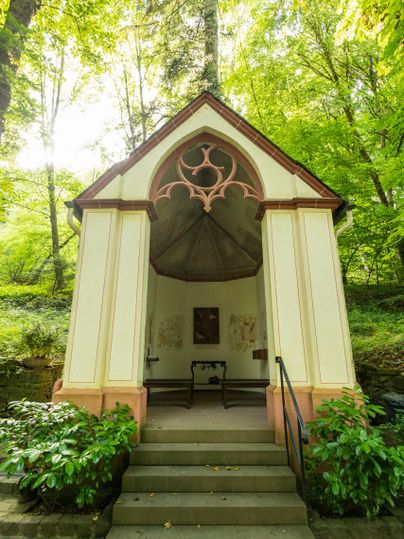 Eine kleine Kapelle steht im Wald, umgeben von hohen Bäumen und grünem Laub. Der Eingang hat eine Treppe und ist farblich ansprechend gestaltet.