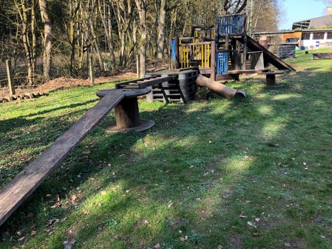 Ein Spielplatz mit Holzstrukturen und verschiedenen Kletterelementen. Die Umgebung ist grün und von Bäumen umgeben.