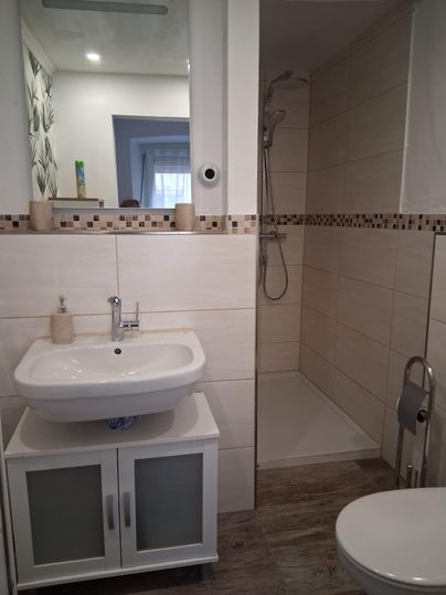 Une salle de bains moderne avec une douche, un lavabo et des toilettes. Les murs sont carrelés en clair et il y a un grand miroir.