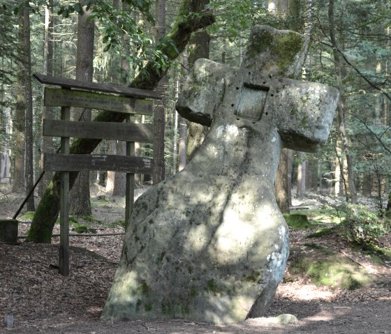 A large, moss-covered stone cross stands in the forest next to a wooden signpost. Sunlight shines through the trees., © Lauschtour.de