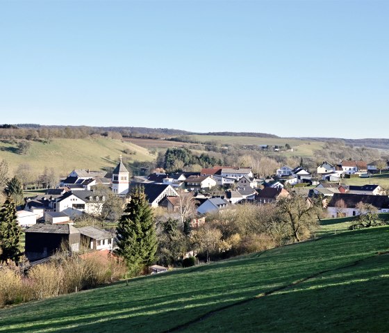 Dorf in der Südeifel mit Kirche, umgeben von grünen Wiesen und Bäumen unter klarem, blauem Himmel., © TI Bitburger Land