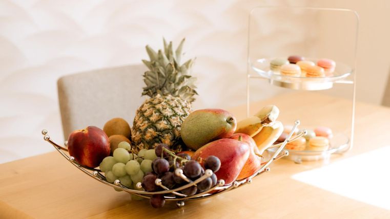 Eine stilvolle Obstschale mit Äpfeln, Trauben, einer Ananas und einer Birne steht auf einem Tisch. Hinter der Schale befindet sich ein Gestell mit Macarons.