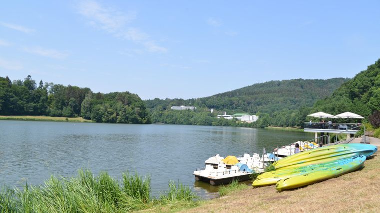 Ein ruhiger See mit einer schönen Uferlandschaft und wachsenden Gräsern. Am Ufer liegen bunte Kajaks und ein Steg mit Freizeitmöglichkeiten.