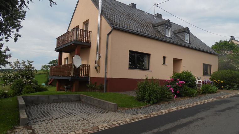 Een eenvoudig woonhuis met een balkon en tuinbloemen. De oprit is geplaveid met stenen en de omgeving is groen.
