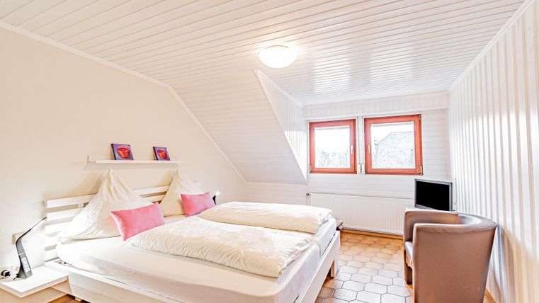 Ein gemütliches Schlafzimmer mit zwei Einzelbetten und rosa Kissen. Es gibt einen modernen Sessel und große Fenster, die viel Licht hereinlassen.