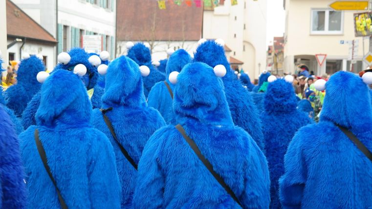 Eine Gruppe von Menschen in blauen Kostümen mit großen Augen, die an ein lustiges Monster erinnern. Sie stehen bei einer festlichen Parade in einer Stadt.
