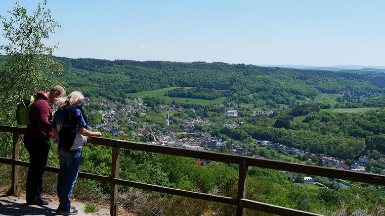 Ein Aussichtspunkt mit Blick auf eine grüne Landschaft und ein Dorf im Tal. Zwei Personen stehen am Geländer und genießen die Aussicht.