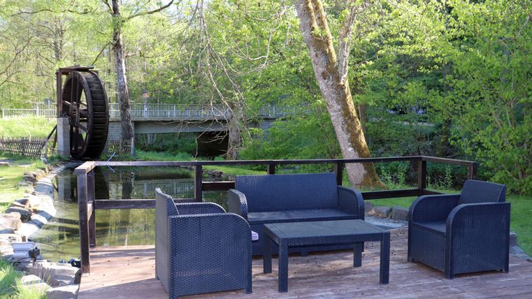 A cozy seating area with rattan furniture is located by a small pond. In the background, there is a water wheel and plenty of greenery.