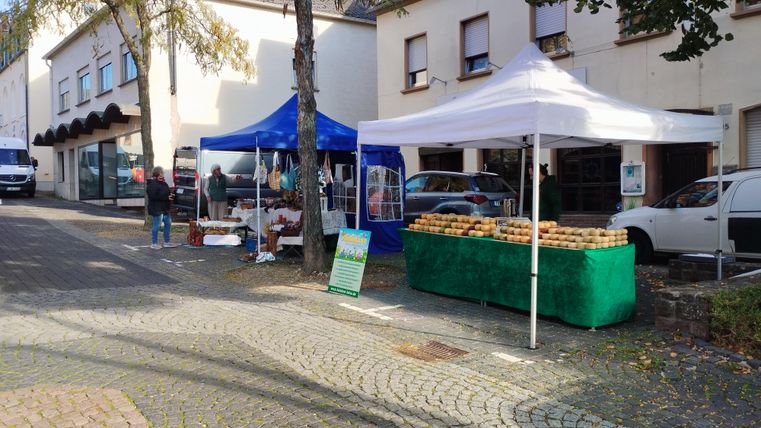 Ein Marktstand mit blauen und weißen Zelten steht an einer Straße. Menschen sind zu sehen, während sie frische Produkte und Lebensmittel kaufen.