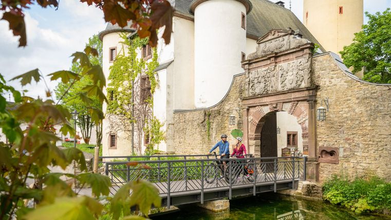 Ein malerisches Schloss mit einer Brücke über einen kleinen Wasserlauf. Zwei Radfahrer genießen die Aussicht inmitten von grüner Natur.