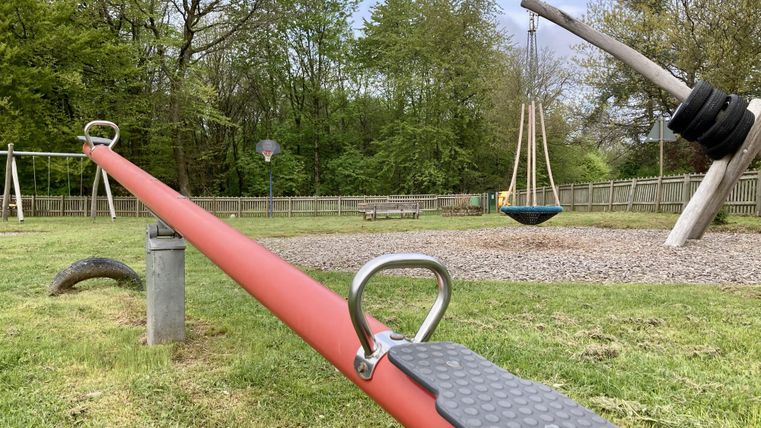 Ein Spielplatz mit einer Wippe und einer Schaukel. Im Hintergrund sind Bäume und eine Rasenfläche zu sehen.