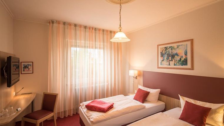 Une chambre confortable avec deux lits simples et un bureau.
De grandes fenêtres avec des rideaux laissent entrer beaucoup de lumière.