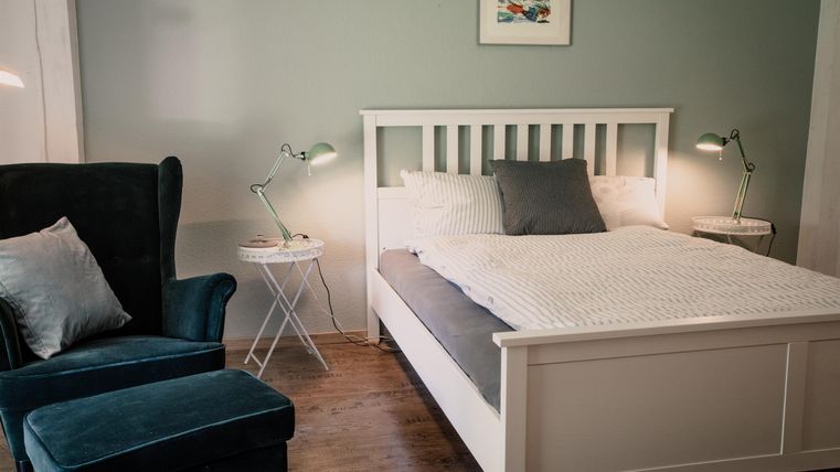 Ein gemütliches Schlafzimmer mit einem weißen Bett und einem blauen Sessel. Die Wände sind in einem sanften Grünton gestrichen, und zwei Nachttische mit Lampen stehen neben dem Bett.