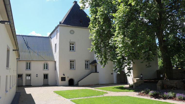 Ein historisches Gebäude mit einem spitzen Dach und angrenzendem grünen Garten. Die Umgebung ist hell und einladend, mit Bäumen und gepflegtem Rasen.