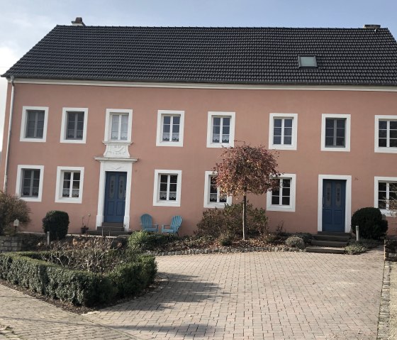 Ein altes Bauernhaus mit rosa Fassade und blauen Türen in Oberweis. Davor stehen zwei blaue Stühle und ein kleiner Baum im Vorgarten., © TI Bitburger Land - Steffi Wagner