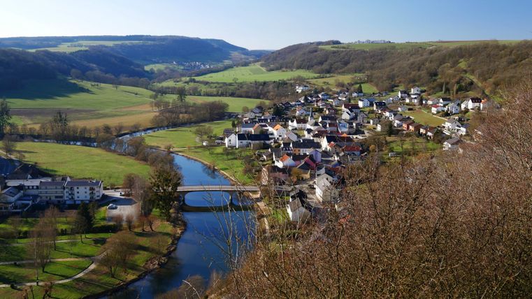 Eine malerische Landschaft mit einem kleinen Dorf am Fluss. Grüne Wiesen und sanfte Hügel umgeben die Szene.