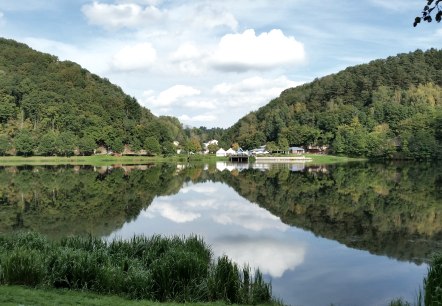 Panorama des Stausee Bitburg mit bewaldeten Hügeln, Gebäuden am Ufer und klarer Spiegelung im Wasser unter blauem Himmel., © TI Bitburger Land