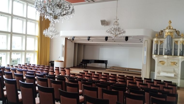 Ein eleganter Veranstaltungsraum mit Reihen von Stühlen und einem Podium. Die Decke ist mit Kronleuchtern geschmückt, und es gibt eine Orgel auf der Bühne.