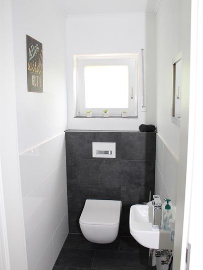 Ein modernes Badezimmer mit einer weißen Toilette und einem kleinen Waschbecken. Die Wände sind teilweise hell und teilweise dunkel gefliest, und es gibt ein Fenster, das natürliches Licht hineinlässt.