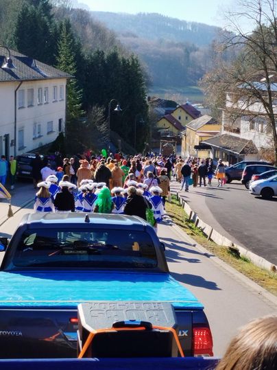 Eine bunte Menschenmenge feiert eine Veranstaltung auf einer Straße. Im Hintergrund sind Häuser und die Natur sichtbar.