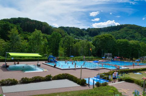 Une piscine avec plusieurs bassins et de nombreuses personnes qui se détendent et nagent. L'environnement est verdoyant et offre de beaux paysages sous un ciel bleu.