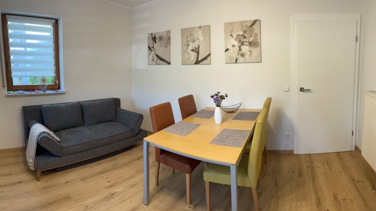 Un salon moderne avec un canapé gris et une table à manger. Des images sont accrochées au mur, et un petit bouquet de fleurs est posé sur la table.