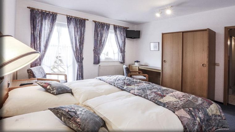 Une chambre confortable avec deux lits simples et un bureau. De grandes fenêtres laissent entrer beaucoup de lumière naturelle, et les rideaux créent une atmosphère agréable.