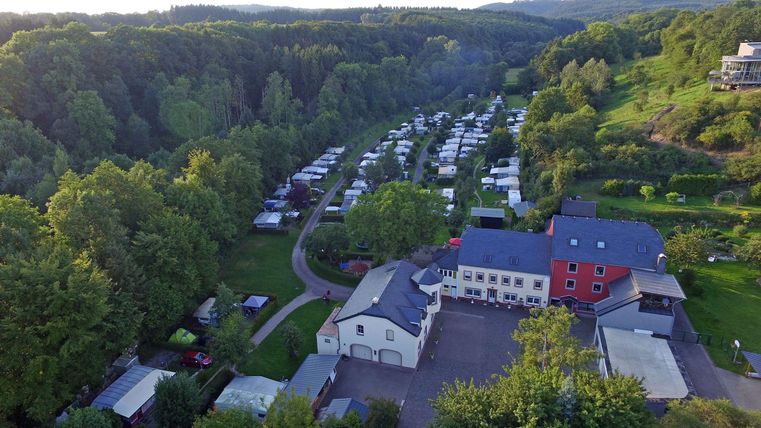 Un camping pittoresque entouré d'arbres verts. En arrière-plan, de nombreux camping-cars et tentes sont visibles.