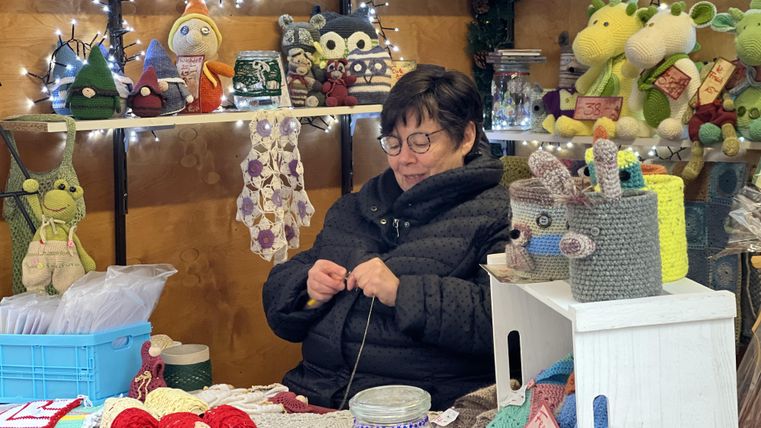 Eine Frau sitzt an einem Tisch und strickt. Im Hintergrund sind farbenfrohe Handarbeiten und Spielzeuge zu sehen.