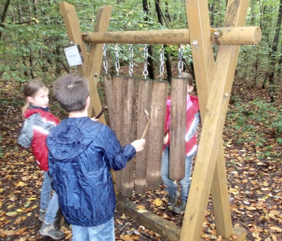 Children play at a wooden sound station in the forest. They are wearing rain jackets and standing on a leaf-covered floor., &copy; Tourist-Information Bitburger Land