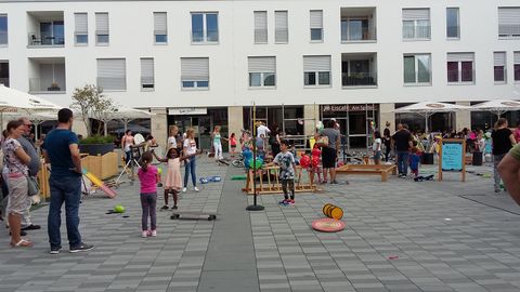 Ein belebter Spielplatz mit Kindern, die verschiedenen Aktivitäten nachgehen. Umgeben von modernem Wohngebäude und Menschen, die zuschauen oder mitmachen.