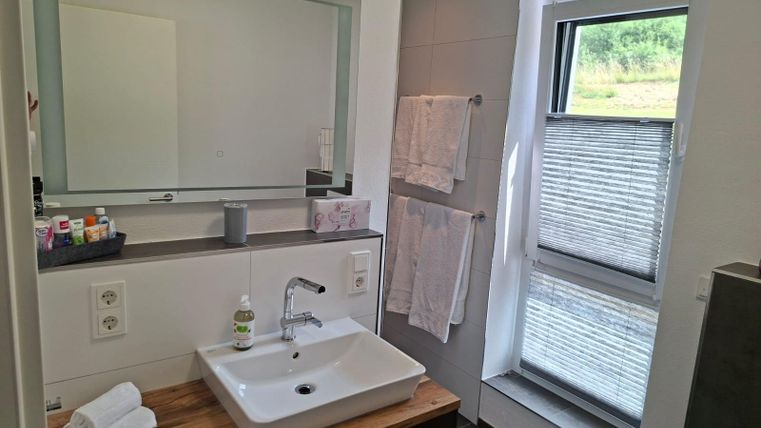 A modern bathroom with a sink on a wooden board and a large mirror. In the background, towels and a window with blinds can be seen.