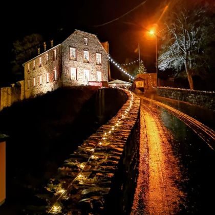 Ein historisches Haus mit beleuchteten Fenstern steht an einem Weg, der sanft ansteigt. Der Weg ist mit Lichtern geschmückt und die Umgebung ist nächtlich und ruhig.