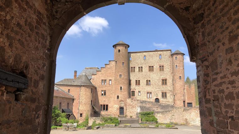 Ein historisches Schloss, gesehen durch ein großes Tor. Der Himmel ist blau und die Landschaft ist grün.