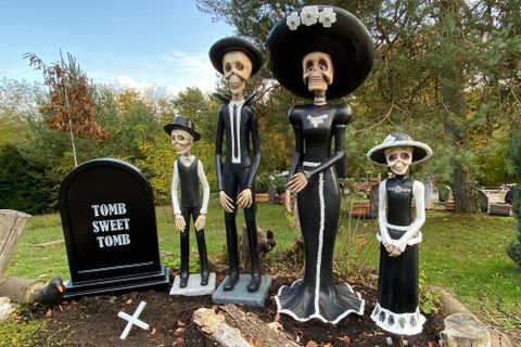 Eine Gruppe von vier Skelettfiguren in festlicher Kleidung steht um einen Grabstein. Der Grabstein trägt die Aufschrift "TOMB SWEET TOMB" und die Umgebung ist mit Pflanzen geschmückt.