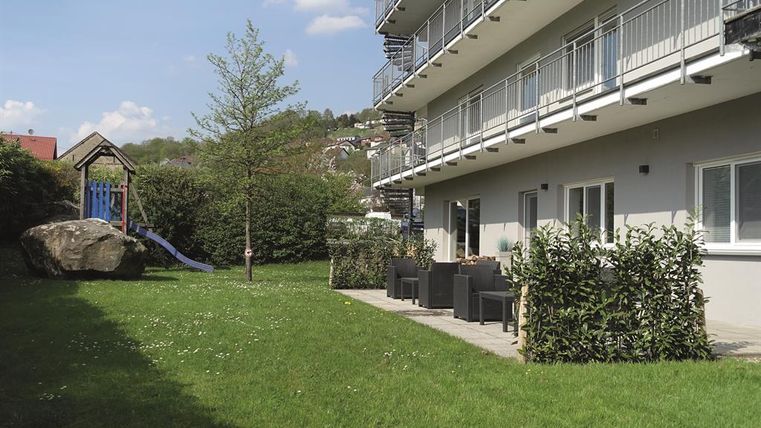 Ein modernes Wohngebäude mit einem grünen Gartenbereich. Im Garten befinden sich Spielgeräte und Sitzgelegenheiten.