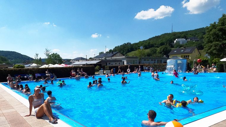 Une piscine pleine de gens par une journée ensoleillée. À l'arrière-plan, des collines vertes et quelques parasols sont visibles.