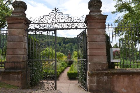 Ein schöner Garten mit einem schmiedeeisernen Tor und üppigem Grün. Im Hintergrund sind Hügel und Bäume sichtbar.