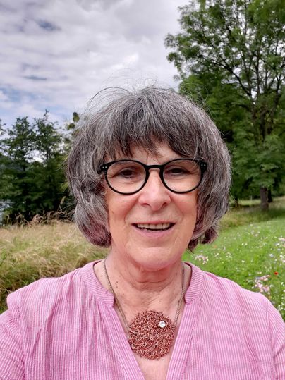 Eine Frau mit grauen Haaren und Brille lächelt in die Kamera. Im Hintergrund sind Bäume und Wiese zu sehen.