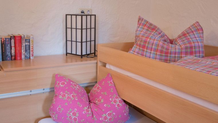 Een gezellige slaapkamer met twee bedden en kleurrijke kussens. Op een nachttafeltje staat een lamp en enkele boeken zijn zichtbaar.