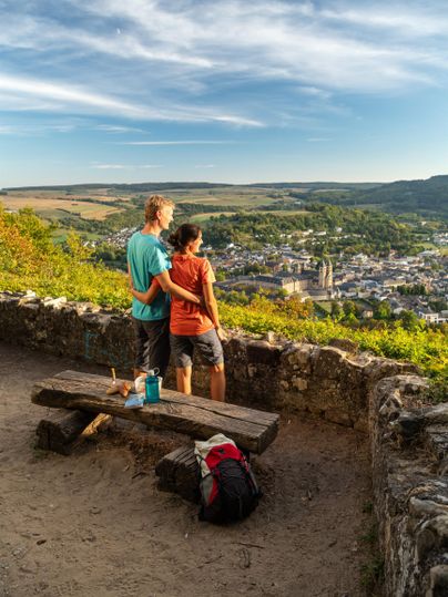Ein Paar steht auf einem Aussichtspunkt und schaut auf die Stadt. Im Vordergrund befindet sich eine Bank, und im Hintergrund sind Hügel und ein klarer Himmel zu sehen.