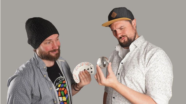 Zwei Männer stehen vor einem grauen Hintergrund. Der links hält ein rundes Objekt und ein weißes Etwas, während der rechts skeptisch auf einen silbernen Gegenstand schaut.